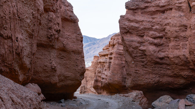 View through the narrow red sandstone walls of Kok-Moinok Canyon, with a rocky dry riverbed trail and distant mountains, Kyrgyzstan, Central Asia