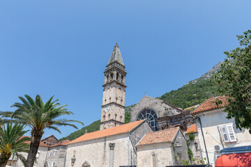 Obraz premium Sunny view of the historic church bell tower and stone buildings with red tile roofs in Perast, Kotor Bay, Montenegro, with green mountains and clear blue sky