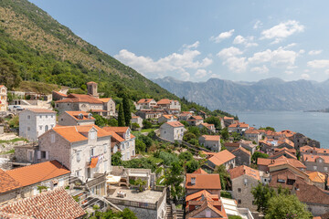 Fototapeta premium Elevated panorama of Perast with stone houses and red tile roofs above the Bay of Kotor, backed by steep mountains and blue sea under a sunny sky, Montenegro