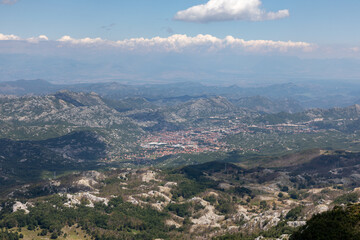 Obraz premium Wide panoramic view from Lovcen National Park overlooking the town of Cetinje, Montenegro, with rugged karst mountains, green forests, distant haze and clouds