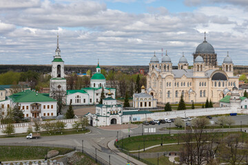 Fototapeta premium Wide cityscape of the Nevyanovsk monastery complex with Orthodox cathedral domes, bell tower and walls, spring trees and dramatic clouds, Russia
