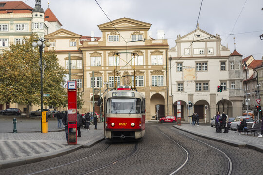 Prague, Czechia - 25 November 2022: View of a classic red and white tram moving along tracks past historic buildings and people at a stop in the city center.