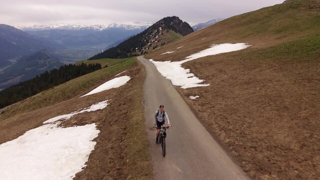 Drone front-facing footage of a mountain biker celebrating at the top of an alpine mountain road, surrounded by panoramic valley views and remaining spring snow patches.