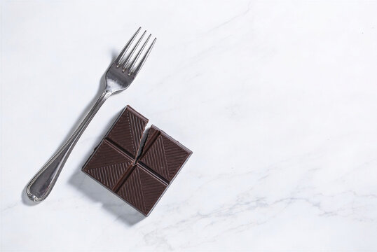 Flat lay of a geometric-patterned dark chocolate bar broken in two pieces next to a vintage silver fork on a white marble background. Stylish World Chocolate Day art. 