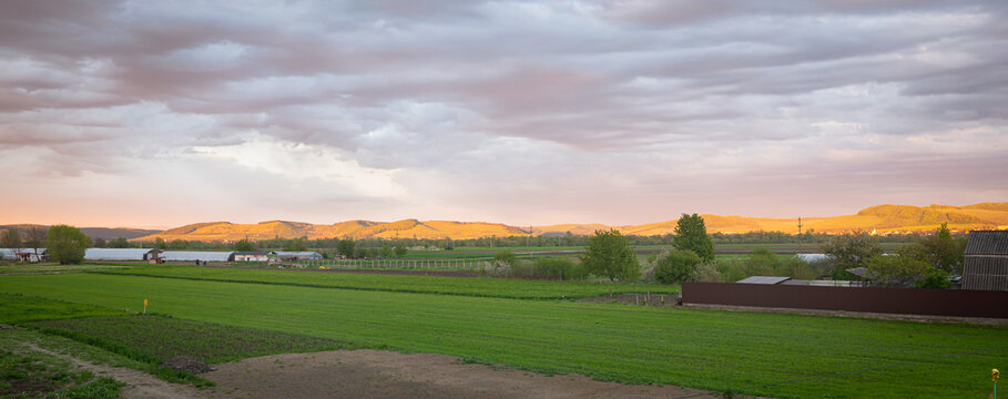 Rural farmland landscape at sunset with rolling hills, green fields and dramatic cloudy sky in Mures valley, central Transylvania, Romania.