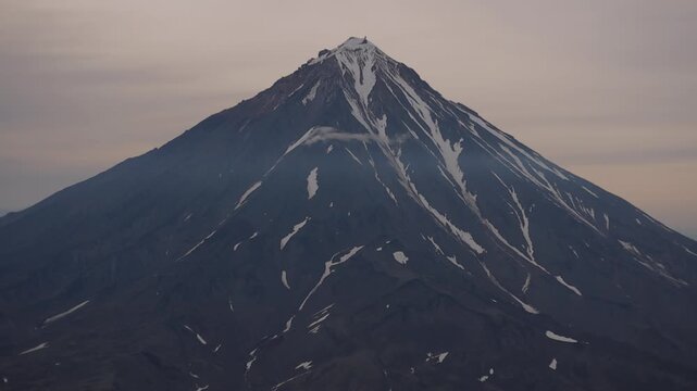 Stunningly beautiful view of majestic volcano with an ice-covered peak and patches of snow between solidified lava against cloudy sky. Concept power of wild nature