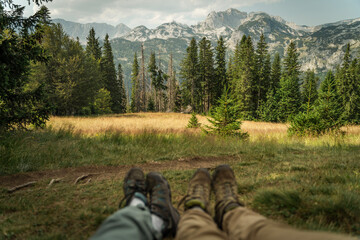 Fototapeta premium Panoramic view of high mountains with blurred legs of hikers in the foreground sitting on a green meadow, selective focus.