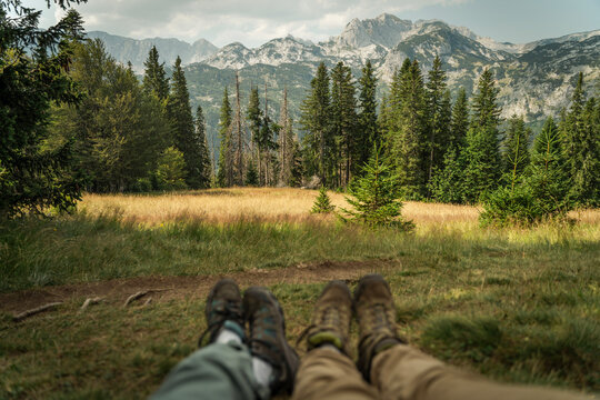 Panoramic view of high mountains with blurred legs of hikers in the foreground sitting on a green meadow, selective focus.