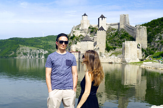Couple standing beside Danube shoreline with Golubac Fortress. Represents emerging Balkan heritage tourism, cross-border cultural discovery, and overlooked Eastern European travel destinations.