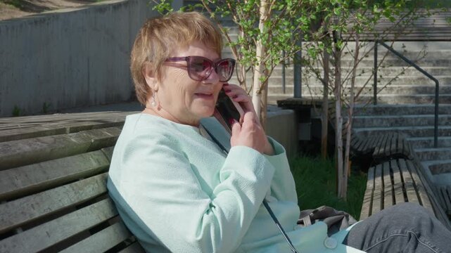 Senior woman talking on smartphone and ending call while sitting on a wooden park bench. Elderly lady chatting on mobile phone outdoors in sunny city. Communication and retirement concept.