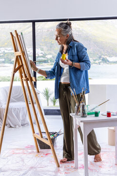 Middle-aged painter painting canvas at easel, holding yellow jar and brush, denim barefoot studio