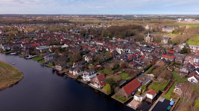 Aerial high angle view of a traditional Dutch town with red roofed houses along a winding river. Drone panorama of a peaceful residential neighborhood and church in the Netherlands countryside.