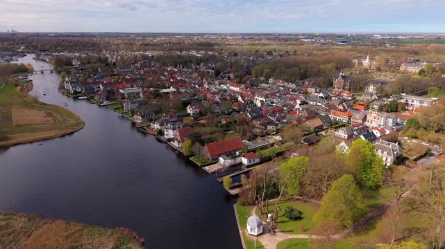 Aerial high angle view of a traditional Dutch town with red roofed houses along a winding river. Drone panorama of a peaceful residential neighborhood and church in the Netherlands countryside.