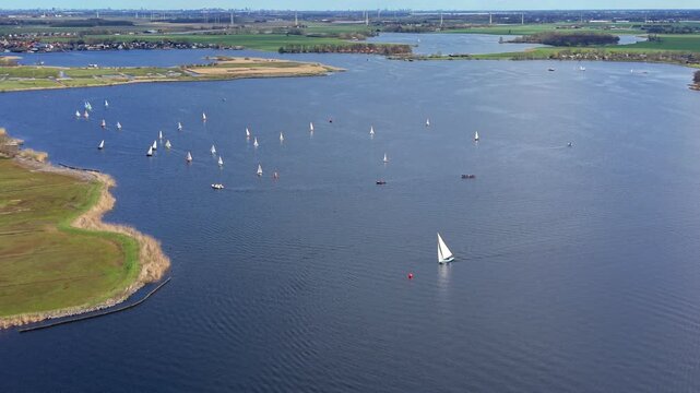 Aerial high angle view of many sailboats racing on a large blue lake in the Netherlands. Scenic landscape of Dutch water recreation, green fields, and distant wind turbines under a clear sky.