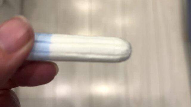 Person holds a tampon in hand, looking closely at its design and colors in a bathroom setting, focusing on product details