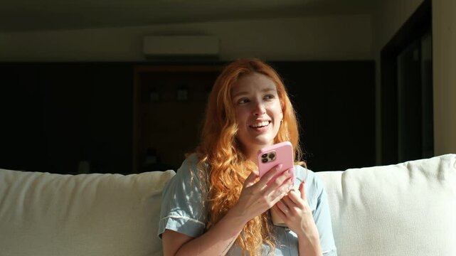 Young woman with beautiful red hair sitting on the couch at home, using her mobile phone to send an audio message while drinking coffee and enjoying the morning sunlight in her living room