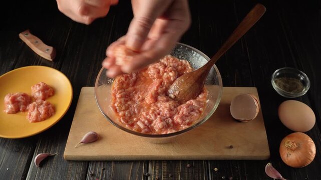 Hands mixing ground meat with spices in a glass bowl, surrounded by garlic, onion, egg, and a yellow plate of meat on a wooden table