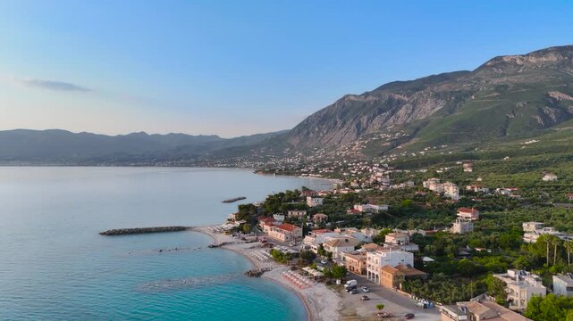 Aerial view of Mikri Mantineia seaside village near Kalamata, Greece, with calm bay, pebble beach, breakwater, coastal houses and green mountain landscape at sunset