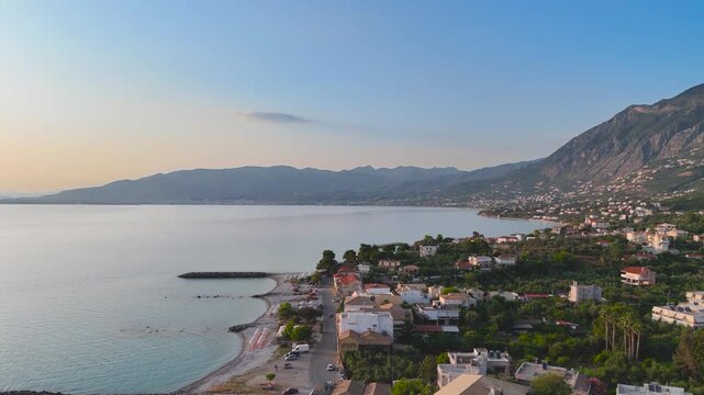Aerial view of Mikri Mantineia coastal village near Kalamata, Greece, with seagulls flying over calm sea, beach, houses, mountains and sunset landscape
