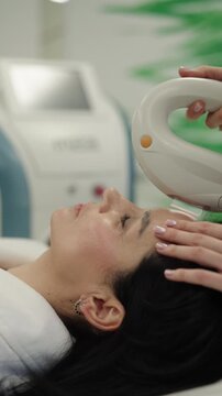 Vertical tracking shot of professional beautician performing non surgical radiofrequency face lifting treatment on female client lying down ina modern aesthetic medicine clinic for skin tightening.