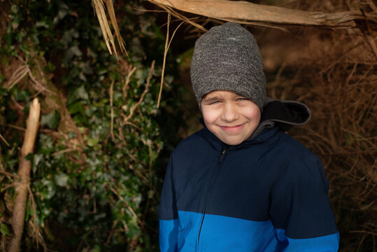 Young boy wearing a gray knit hat and blue winter jacket smiles while standing outdoors. Boy resting after running in forest with his father