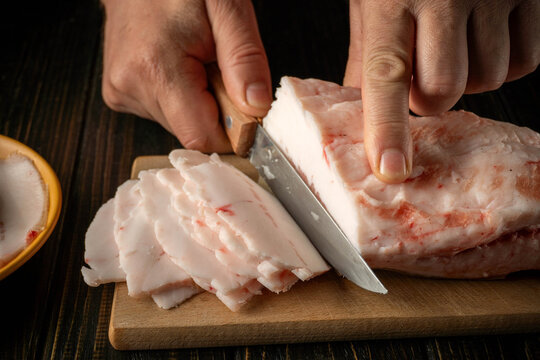 Hands holding a knife, cutting a piece of pork fat. The pieces are carefully placed on a wooden board. A small bowl is on the table nearby