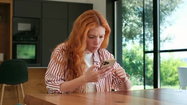 Annoyed redhead woman at home holding a mobile phone, reacting with anger and sadness to a message, feeling stressed and upset after reading negative news on her social media feed