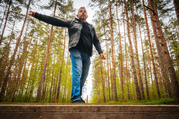 Fototapeta premium Male figure in black jacket and blue jeans stands on wooden platform, arms extended, surrounded by tall pine trees in a lush forest setting