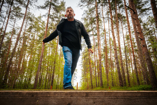 Middle-aged man walking along a wooden path in a dense forest, surrounded by tall pine trees and lush greenery, with a clear sky visible above