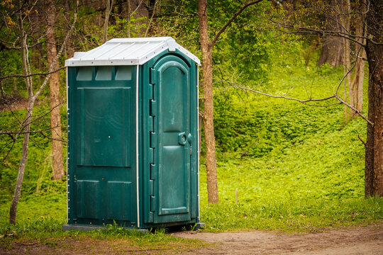Green portable toilet structure positioned on a dirt path surrounded by lush greenery and trees in a natural outdoor setting