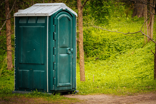 Green portable toilet structure positioned on a dirt path surrounded by lush greenery and trees in a natural outdoor setting