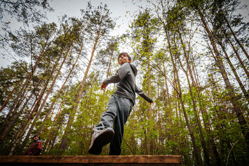 Fototapeta premium Young male wearing gray hoodie and sweatpants walks on a wooden platform in a forest, with tall trees and greenery surrounding the scene