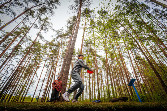 Young male disc golfer in gray sweatshirt throws disc towards target while female companion watches in a forested area with tall trees and green foliage