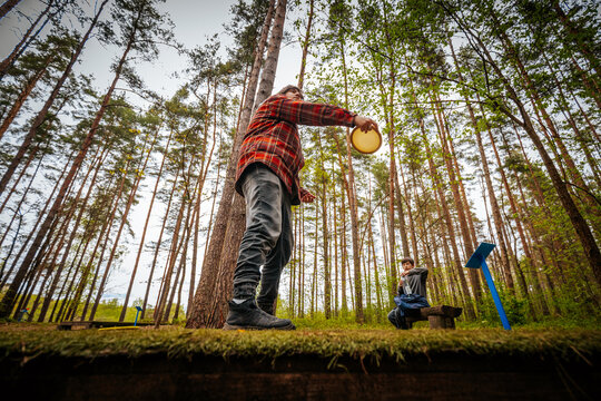 Male disc golfer in red plaid shirt prepares to throw disc towards target while female player sits on bench in lush green forest setting with tall trees