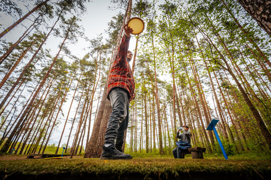 Man in red plaid shirt holds a round object above his head while a woman sits nearby in a forested area with tall trees and green foliage visible