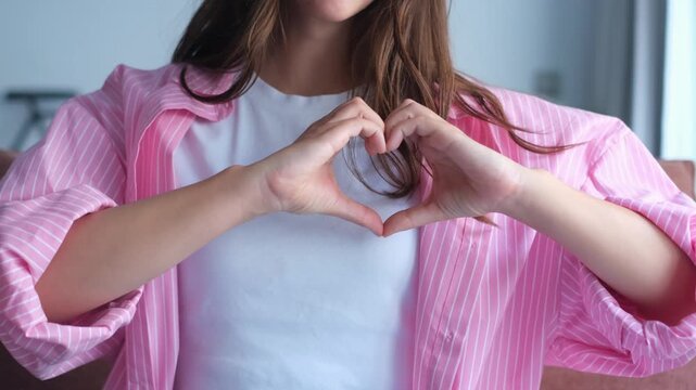 Closeup of a smiling young woman in a pink shirt making a heart shape with her hands, expressing love, gratitude, and support while showing a gesture of affection and kindness