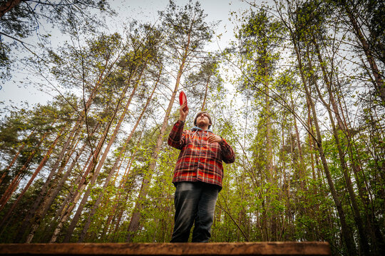 Male wearing plaid shirt stands on wooden platform in forest, holding red object while surrounded by tall trees and green foliage under a cloudy sky