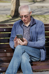 Obraz premium An elderly pensioner sits on a park bench and looks at his smartphone.