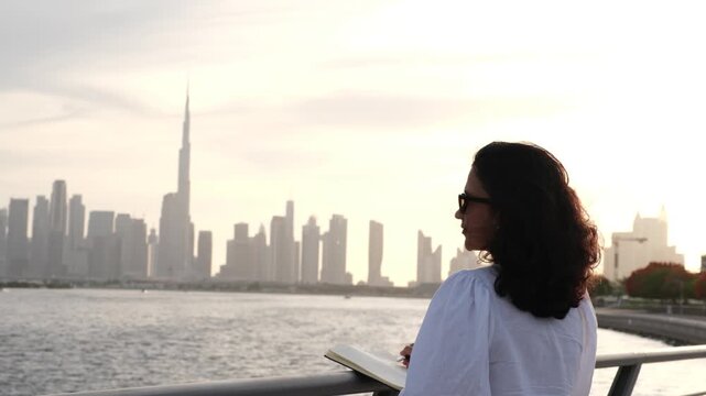 woman writing in notebook by Dubai waterfront with peaceful skyline sunset video in 4k resolution