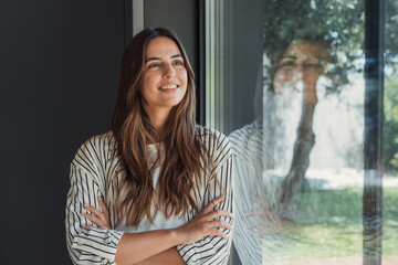 Fototapeta premium Cheerful young woman standing indoors with arms crossed, smiling as she looks out a large window, reflecting a blurred garden with trees and natural daylight