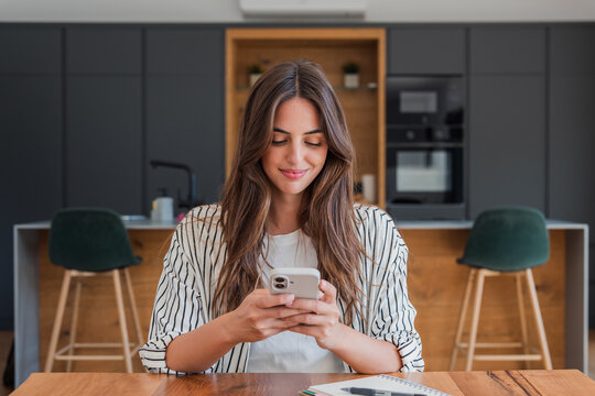 Young woman sitting at a table in a stylish contemporary kitchen, smiling and engaging with her mobile phone, using social media or communication apps