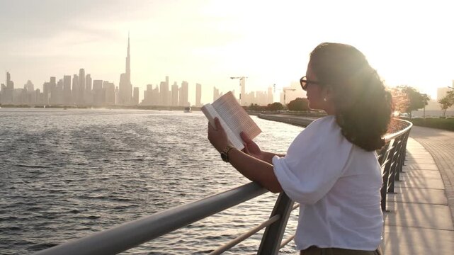 Woman reading book by Dubai waterfront during sunset with peaceful lifestyle atmosphere