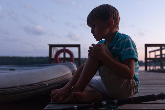 Peaceful child sits on a weathered dock angling in a serene lake. Youngster waits patiently with fishing rod by the calm water. Kid enjoys quiet outdoor relaxation in rustic natural scenery