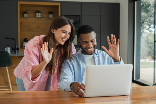 Happy ethnically diverse couple waving to the camera during a video call on a laptop, connecting and communicating online from their modern home kitchen