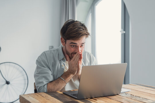 Young man reacting to surprising news or unexpected information on his laptop, expressing disbelief and wonder while sitting at a wooden desk in a bright room