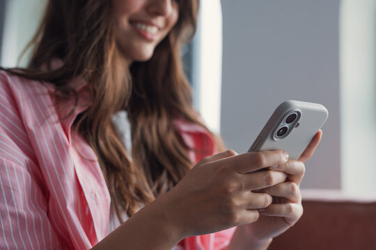 Woman holding a mobile phone in both hands, smiling, and feeling happy while texting, browsing the internet, or engaging with social media applications