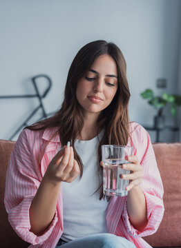 Young woman holding a white pill and glass of water, about to take daily medication at home for health, wellness and self-care routine in a casual living room setting