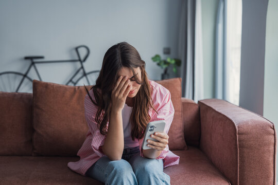 Young woman sitting on a sofa at home, crying and covering her face with one hand while holding a mobile phone in the other, expressing stress, sadness, and emotional distress over bad news