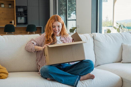 Red-haired woman expresses joy and satisfaction while opening a new online shopping delivery package on her living room couch at home, enjoying her recent purchase