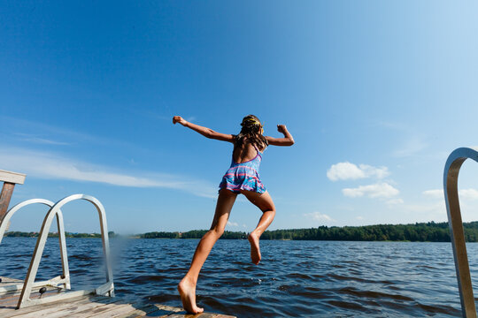 Girl jumps off a dock into a lake creating a splash on a sunny summer day. Child in swimsuit leaping with joyful energy. Kid experiences carefree outdoor fun and refreshing water recreation